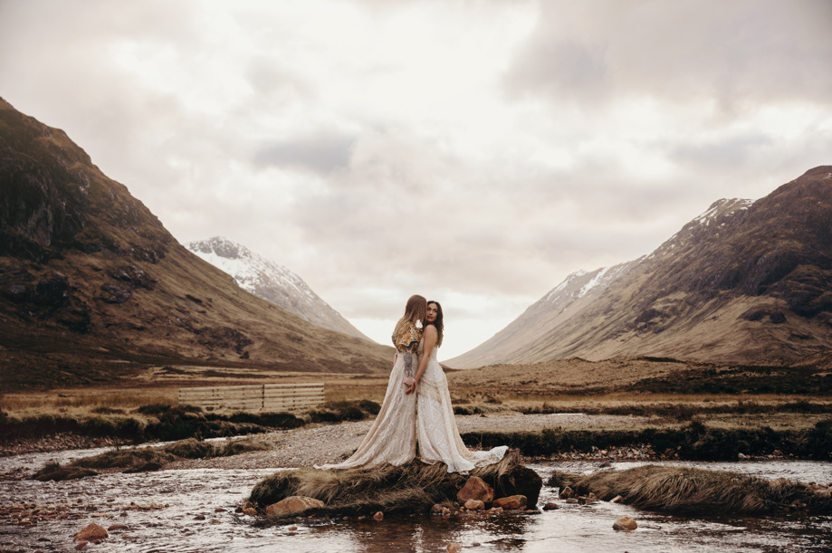 Couple holding hands on a small river island in Glencoe, Scottish Highlands wedding setting with mountains reflected in water